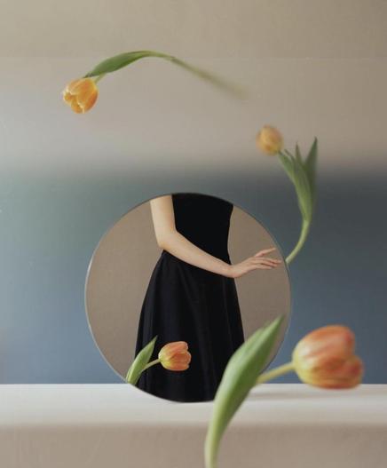Photography. A color photo of a young woman as a reflection in a round mirror and several carnation flowers.  A rimless round mirror stands on a white border. A blue room wall can be seen in the background. The reflection shows the arm of a young woman and her black dress. The head is not visible. In the foreground is an orange tulip, which can also be seen in the reflection. Two tulips are in motion and circle the mirror in the air.