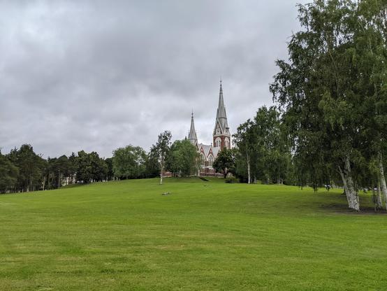 L'église de Joensuu au fond au centre, au premier plan une pelouse d'un parc, au fond des arbres et au-dessus un ciel gris