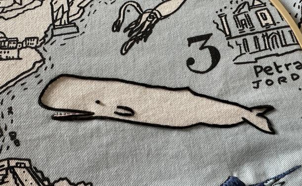 A sperm whale on a printed fabric map. The whale is white and it has been outlined in black stem stitch