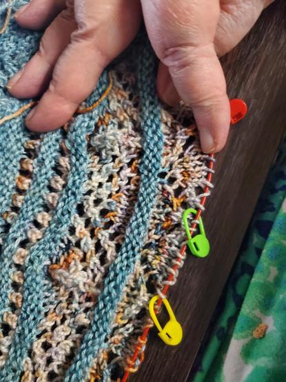 My hand showing the 2 color lace shawl while on needles with brightly colored plastic pins denoting each of 44 sections repeating, the Clue #3 section