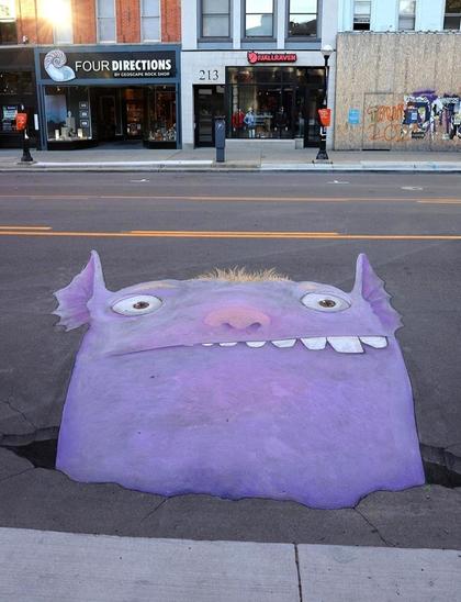 Streetart. On one side of the street, a purple monster (called Norman) has been drawn in chalk. The unusually big and fuzzy guy has a big pink nose, small ears and crooked teeth, of which you can see some big, round teeth on the right. He looks out of a (painted) hole in the road and looks a bit intimidated
Title: 