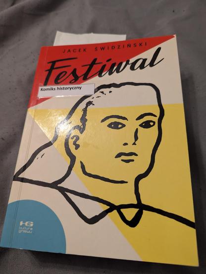 Okładka książki Festiwal — rysy osoby czarną kreską, na błękitno czerwono żółtym abstrakcyjnym tle. Nalepka biblioteczna Komiks historyczny