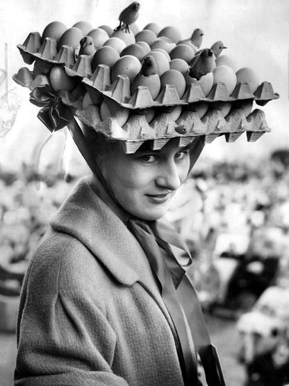 black & white photo of woman wearing open, square carton of eggs tied to the top of her head with a ribbon — photographer unknown