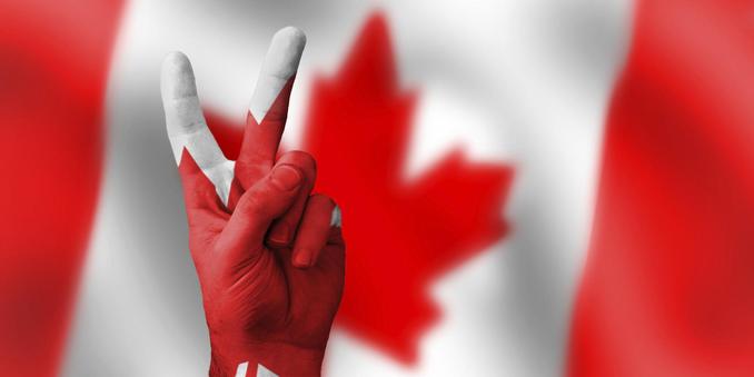 Hand making a peace symbol painted with the Canadian flag in front of a Canadian flag. 