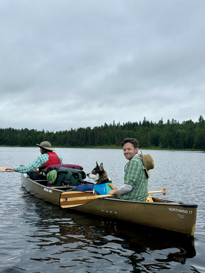 Two people and a dog in a canoe loaded with several packs paddling on a lake surrounded by coniferous forest