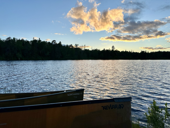 Clouds and tree line at sunset viewed from across a lake with two beached canoes in foreground