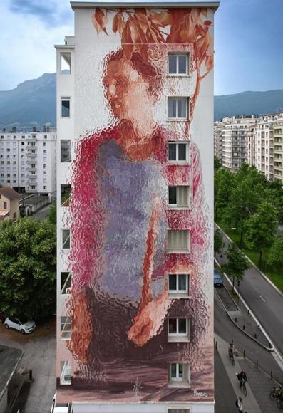 Streetartwall. A huge mural of a young man has been painted over 7 floors on the outside wall of a narrow high-rise building. The brown-haired man is only vaguely recognizable. It looks as if he is standing behind frosted glass - the figure can only be guessed at. A standing man in brown trousers and a gray-pink shirt stands with a thin, pink tree and looks into the distance. Fantastic.
Info: The painting style of the blurred krakelee is painted with a brush and is the trademark of the artist Fintan Magee.