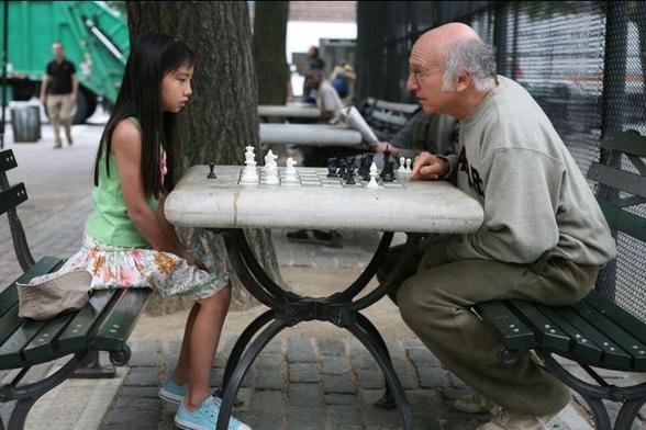 In a scene from Whatever Works, Larry David leans over an outdoor chessboard toward his opponent, Willa Cuthrell-Tuttleman.