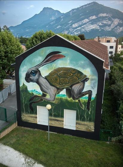 Streetartwall. The mural of a unique animal was sprayed/painted on the two-storey exterior wall of an elementary school with two windows. A hare with long ears and legs and a tortoise shell on its back hops through a green landscape of trees, meadows and hills. This unusual animal is depicted in rich detail in shades of blue, gray and green. (The photo shows a green meadow in front of the mural and the imposing gray mountains behind it.)
Info: 