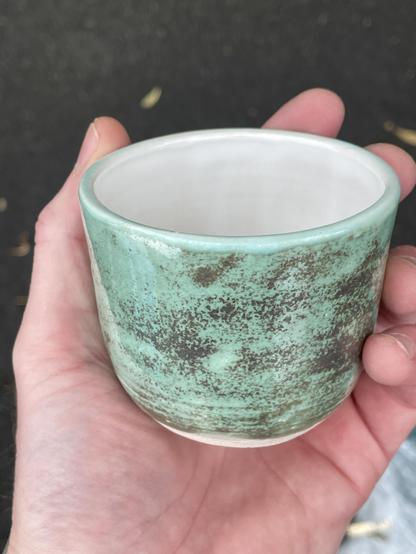 A small beaker style pot, held in my hand. It is glazed a copper-green on the outside with metallic copper oxide patterning, and a glossy white interior.