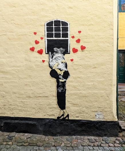 Streetartwall. A magnificent mural depicting a couple in love was sprayed onto a yellow house wall. The mural is stencil art and is painted in red, black and white. You can see a small window from which a young man is hanging and kissing his girlfriend, who is standing under the window. Their heads are right next to each other and the young woman is hugging the man's back. There are many red hearts around the window.
Info: Tabby spray-painted this mural in Buffalo back in 2022. The advantage of stencil art is that you can work quickly and reproduce it in different places and on posters.