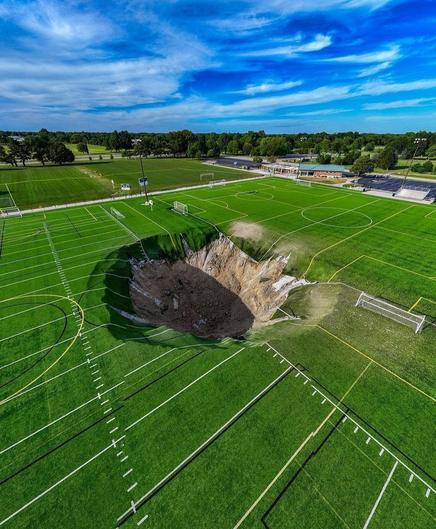 Photography. A color photo of a soccer pitch with a round, gigantic hole in the middle of the pitch. It looks surreal, the lines of the field now lead into the round hole. Blue sky, forest and another soccer pitch can be seen in the background. The whole thing looks like something out of a Hollywood sci-fi movie.
Info: An active limestone mine has collapsed under Gordon F. Moore Community Park in Alton, IL. The whole thing happened a few days after the end of a soccer camp for children. A perfectly round hole opened up and swallowed one of the field lights. The hole is estimated to be 100 feet wide and 30 feet deep, although the mine is much deeper. A surveillance camera even captured a video of it. The entire park is currently closed.