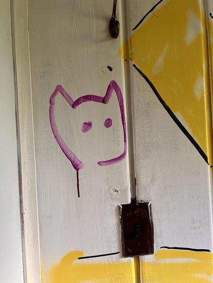 Graffiti of a simple, purple cat face drawn on a white wooden door with yellow paint nearby and a dark brown light switch below. A spoon is hanging on the top of the door.