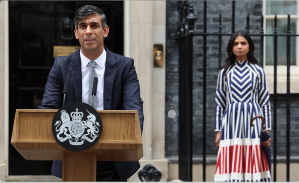 A failed tax evading billionaire’s wife stands behind him looking glum wearing a dress that appears to have been made up out of the waste from many hideous clown costumes.
