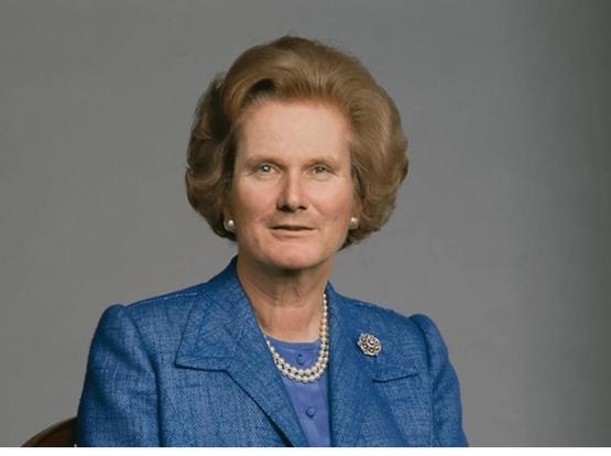 Elderly woman with styled hair, wearing a blue suit, pearls, and a brooch, posed against a grey background. Yes is Kier Thatcher.