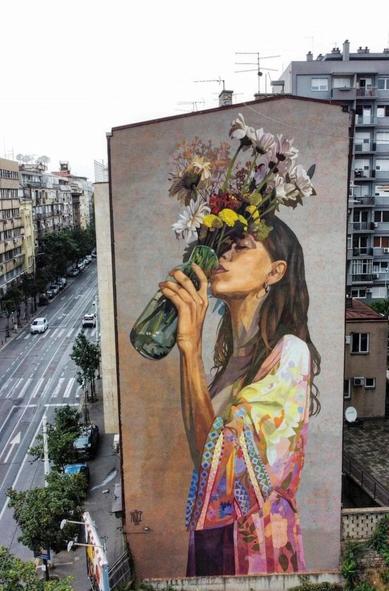 Streetartwall. A beautiful mural of a young woman with a vase of flowers was sprayed/painted on the outside of a five-storey residential building. The portrait shows a girl with long brown hair in a pastel-colored, colorful silk coat. She is drinking water from a glass vase that is abundantly filled with colorful flowers. The mural forms a wonderful contrast to the gray city around it. The girl is striving for the energy that only nature can give us.
