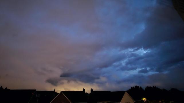 Here's a long description of cloudy night sky with rooftops along the bottom of the frame. The sky is various shades of blue with cloud edges to the right, as that's where the light was creeping in for a later sunrise. Low centre is a patch of yellow light you would think is lightning but is actually the glow of lights from a town 20 miles away. Above it is what looks like the top half of a dragon head which gives the photo it's slightly menacing look. The left half of the sky is orange/light brown with some light blue cloud patches
