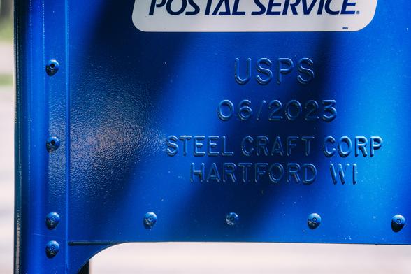A blue post office mailbox with letters stamped in Gorton: USPS 06/2023 STEELCRAFT CORP HARTFORD WI