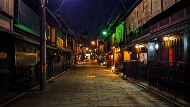 Nighttime shot of empty stone paved street with 2 storey houses on either side with no gaps in between. The houses have wooden fronts and window slats and upper storey with thin rolling blinds. Street lamps with different colours and some yellow lamps over some doors illuminate the street. 

#Gion #Kyoto #Japan #Travel #Photography #StreetPhotography 
Pic from Dec 2023