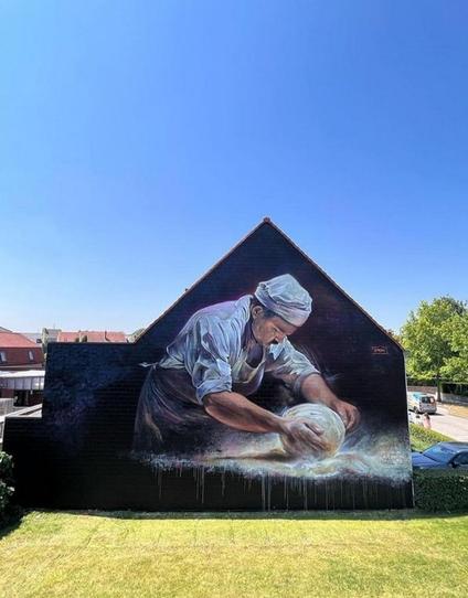 Streetartwall. On the brick wall of a two-storey bakery building, a beautiful mural of a baker kneading dough has been painted in the style of old  Flemish masters oil paintings. The background is black and brown, allowing the man in his white baker's clothes to take center stage. He is depicted from the side and is concentrating entirely on kneading a large round lump of dough in front of him. You can almost see the movement of rolling and kneading in the light-colored flour. A beautifully designed mural in the right place. Masterpiece. (The photo shows a meadow in front of the house and houses in the surrounding area)