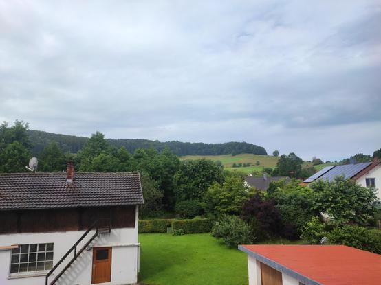 Blick vom Balkon auf eine leichte Anhöhung mit Blick auf grünen Wiesen und Wald.
Im Vordergrund eine alte Schreinerei.
Wolken am Himmel und es regnet....
Es wird schwül.....