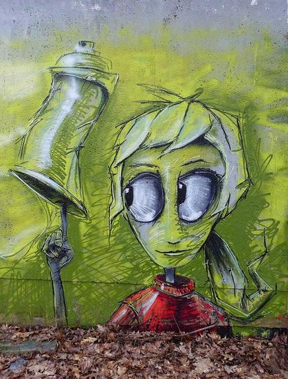 Streetartwall. A cute mural with a green creature and a can of spray paint was sprayed/painted on a hidden wall in the forest. It looks like a little green girl with very big eyes, a ponytail hairstyle and a red sweater. It is depicted near the ground and from the shoulders up, balancing a dented spray can on a thin, long finger while looking at it with big eyes.