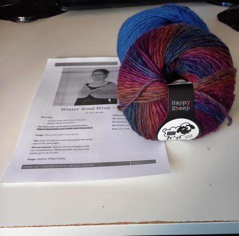 A pattern printout and two skeins of wool on a white desk. One skein is blue with a slight purple tone to it. The othed is multicolored ranging from brownish through deep winered  to blue.