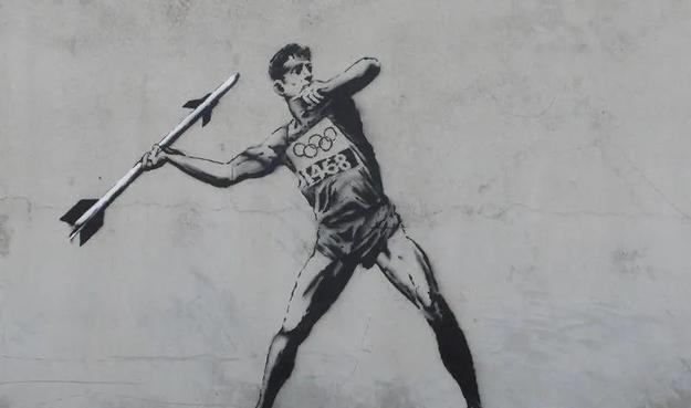 Stencil graffiti of an athlete wearing Olympic gear, poised to throw a javelin that is actually a missile, on a gray wall.
