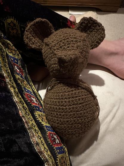 A small brown crocheted bear without arms, legs or facial features leaning against multicoloured velour trousers. 