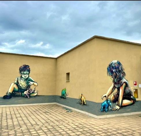 Streetartwall. Two small children playing with mammoth figures were sprayed/painted on the yellow exterior walls of a modern low-rise building (a museum). The detailed and finely drawn mural shows a girl sitting on the floor with a blue mini mammoth in the foreground, then two more mammoths in green and yellow on the long side and a little boy with glasses playing with a pink mammoth on the adjacent wall. A wonderful idea for the entrance to a paleontological museum.