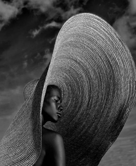 Photography. A black and white photo of a woman wearing a large sun hat. The black woman is photographed from the side. You can only see her face and one upper arm. The background is a cloudy sky. But the large straw hat dominates the picture. It is draped upright, framing the young woman and making the photo appear dramatic in black and white.
