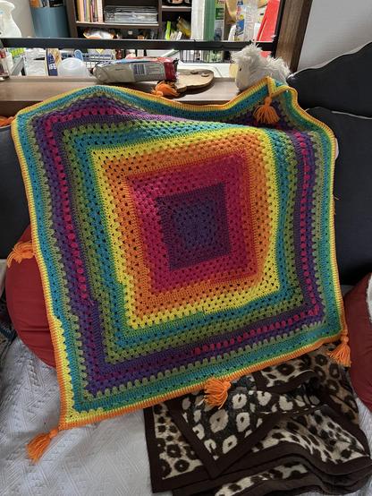 A large square crocheted blanket with a radial colour change! The colours go dark blue, purple, pink, orange, yellow, light blue, green, purple and pink, green, light blue, yellow and orange as you move from the middle outwards. There are 8 golden tassels around the outside.
