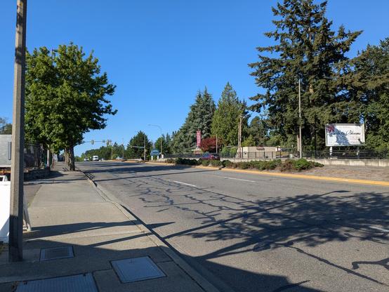 A view of SR-99 outside a coffee hut. The street has wide, empty sidewalks, a few street trees, and mostly empty wide lanes for traffic. There are two lanes in each direction separated by a raised median. No crosswalks are visible.