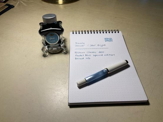 Ink swatch on an A5 Rhodia dot pad. On the top left is a Diamine Glacier ink bottle, made of glass with four legs. At the bottom right is a pastel blue and white Pelikan Classic fountain pen.
