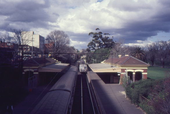 A view taken from an overbridge looking down onto a railway station with two tracks running between side platforms.  On each platform stands a polychrome brick station building, each with an awning projecting to the platform edge.  We can see the roof of an electric train standing at the platform at left, and another train is approaching the station on the other track at right.