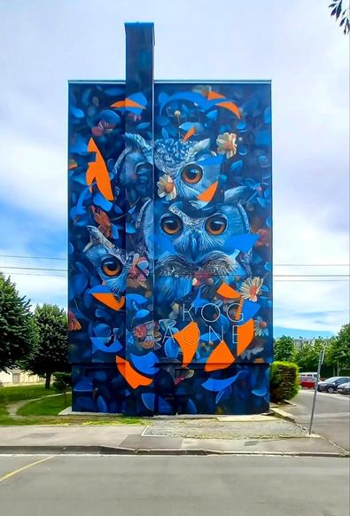 Streetartwall. The impressive mural of a blue owl among blue leaves and flowers was sprayed/painted on the exterior wall of a four-story modern building. The main colors are shades of blue, orange and white. The surface is covered with small blue leaves and flowers, interrupted by small orange arc-shaped splinters. In between are the large brown eyes of an owl, whose head is repeated several times on the wall. The artist 