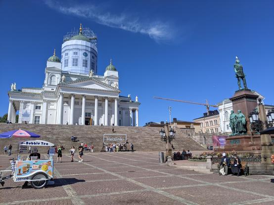 La cathédrale de Helsinki, blanche avec des dômes verts surmontés de croix dorées. Au premier plan à droite une statue, une mouette est posée sur la tête du personnage. Au premier plan à gauche un stand de Bubble Tea. Des escaliers imposants mènent à la cathédrale. Le ciel est bleu, avec juste un tout petit nuage blanc qui passe.