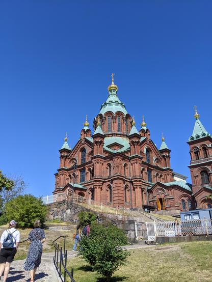La cathédrale orthodoxe de Helsinki, en briques rouges, toiture verte et sommets dorés, sur fond de ciel parfaitement bleu. Elle est au sommet d'une colline, au premier plan un escalier qui monte et un petit parc.