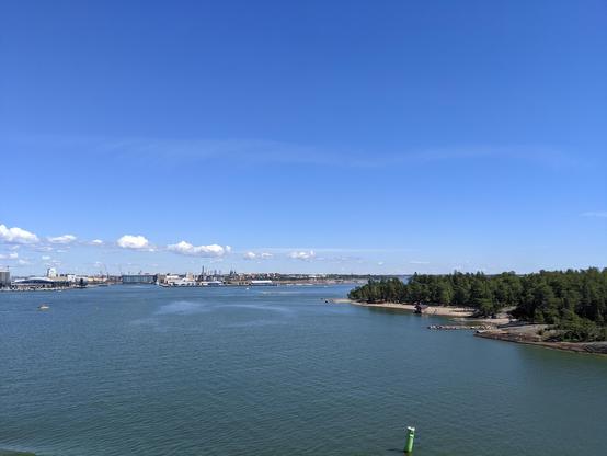 La ville de Helsinki au loin vue depuis le ferry en pleine mer. Au premier plan à droite, une île boisée. Le ciel est bleu avec à peine quelques nuages.