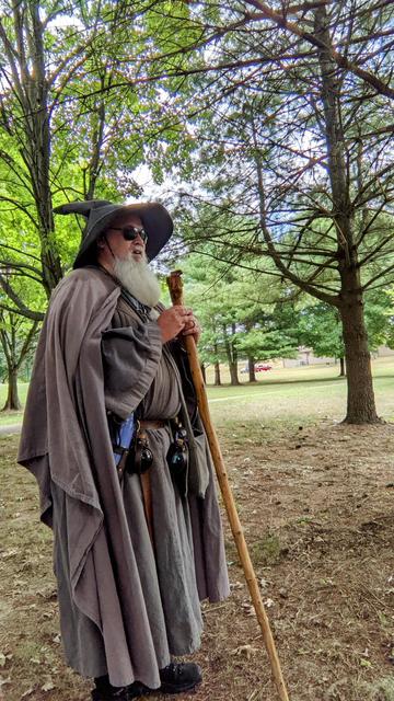 middle aged man with grey beard and Gandalf style wizard outfit with hat, staff, & sunglasses standing in a grove of trees