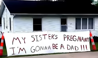 A large white hand painted sign sits on the ground in front of a wooden bungalow building. In large untidy letters the sign reads 'MY SISTER'S PREGNANT I'M GONNA BE A DAD !!!