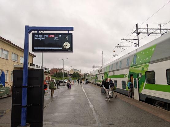 Le quai de la gare de Rovaniemi. Au premier plan à gauche, le panneau indiquant la destination (Kemijärvi), l'horaire (7h35 retardé à 7h51), le numéro du train (PYO 265), les gares desservies (Misi, Kemijärvi), une horloge etc.

À droite le train couchettes à deux niveaux, vert gris et blanc. Le ciel est gris, il pleut. Des gens sur le quai sortent du train.