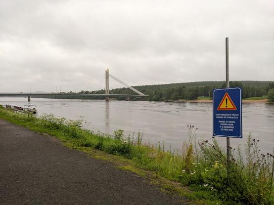 La rivière Kemijoki, extrêmement large. Au fond à gauche, un pont suspendu enjambe la rivière. Au premier plan à droite, un panneau indiquant qu'il ne faut pas se baigner ici car le courant est fort.
