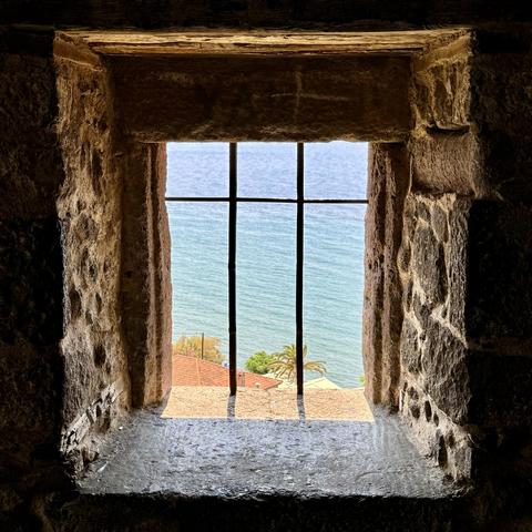A fortress window with iron fence. Behind it the sea is visible. At the bottom of the visible area there are a few roofs and a few tops of trees.