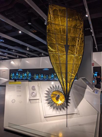 Photograph of a museum exhibit, showing a gold petal hanging from the ceiling, a sunflower shaped Starshade model in a case, and two spiral origamis in a case