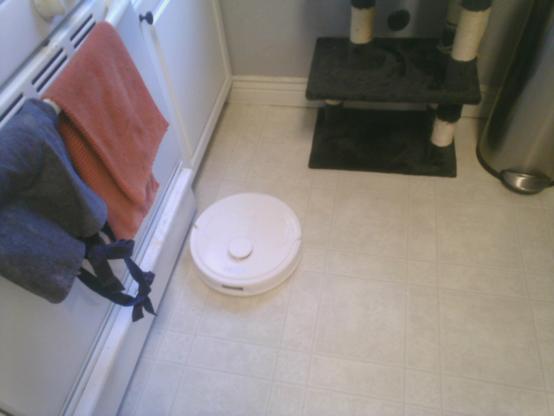 A photo of Rosie the robot vacuum mopping a kitchen floor. Towels and an apron hanging from an oven door are visible on the left side of the photo. In the top right the bottom of a small cat tree and a metal trash can are visible