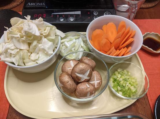 A tray with bowls with cut or sliced vegetables (cabbage, scallions, carrots) and one with mushrooms with tops scored