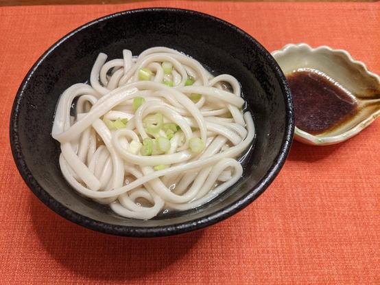 A small bowl with udon and some dashi and scallions on top with a small dish of soy next to it
