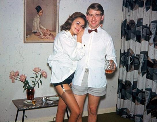 Old photo of two people wearing semi formal tops but no bottoms, just underwear, smiling for the camera. One is holding a drink. 