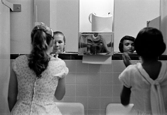 Black and white photo of a young white woman and young Black woman in a public restroom standing next to each other at a mirror and applying makeup